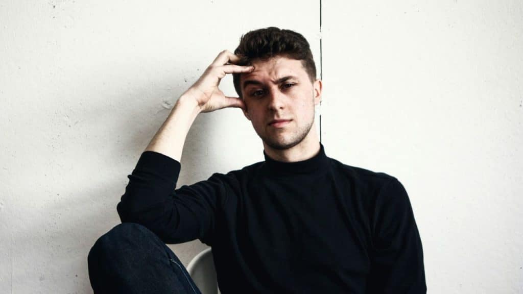 A man in a black turtleneck rests his head on his hand and looks at the camera.