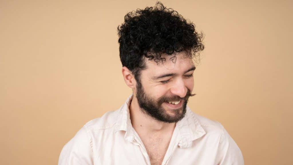 A man with a mustache and curly hair smiles and looks down.