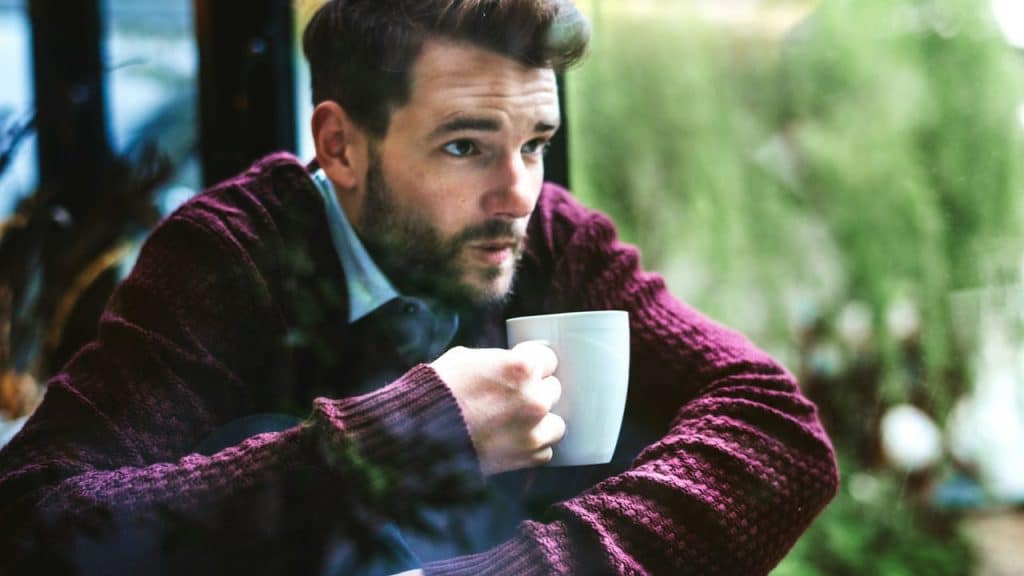 A man with a beard sits indoors, holding a white mug and looking out a window.