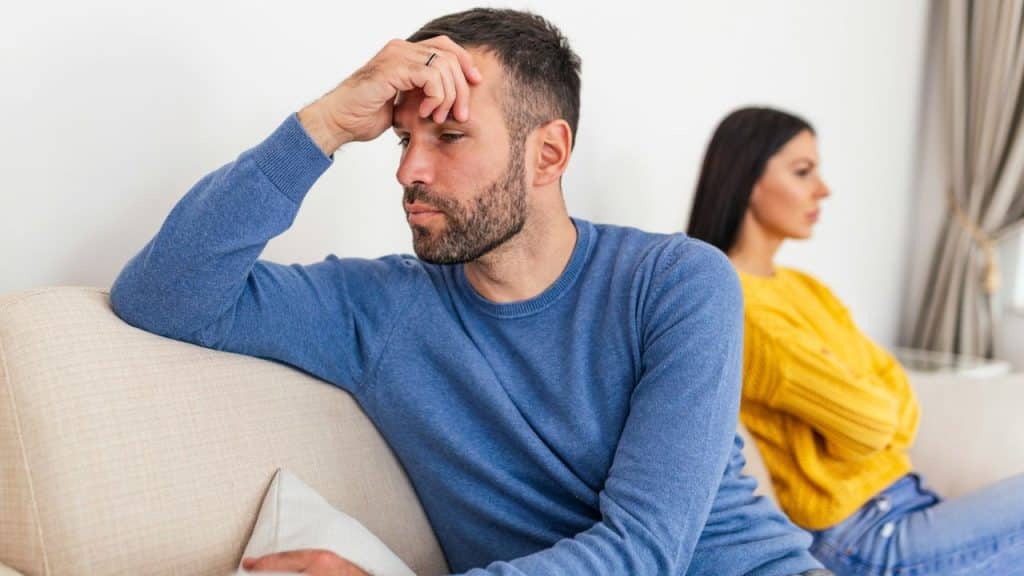 A man and woman sit on a sofa, facing away from each other with frustrated expressions.