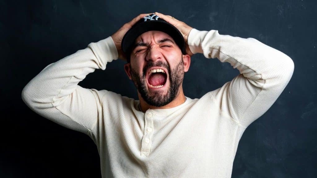 A man with a beard and a baseball cap clutches his head and screams.