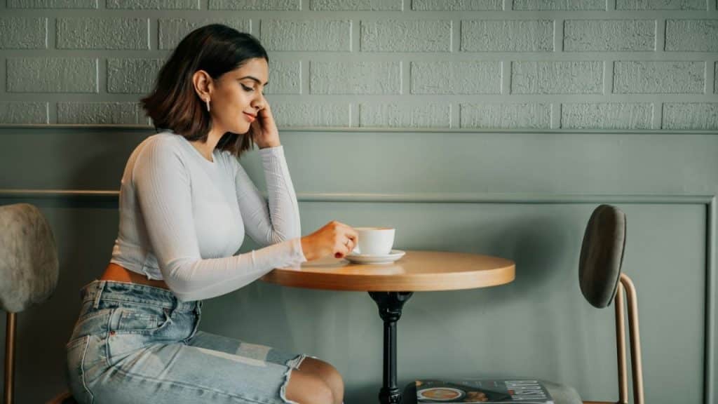 A woman with a short haircut sits alone at a coffee table, holding a cup and saucer.