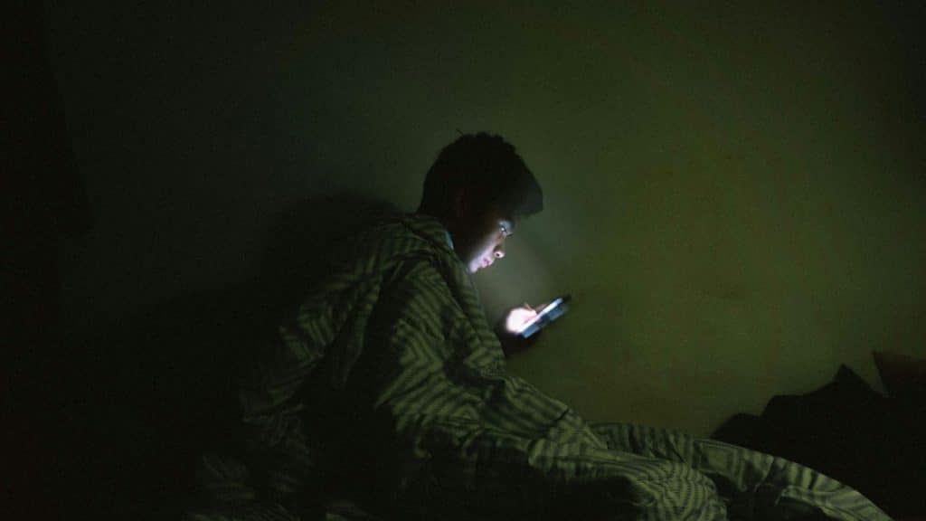 A person sits in a dark room under a blanket, looking at a phone with a glowing screen.