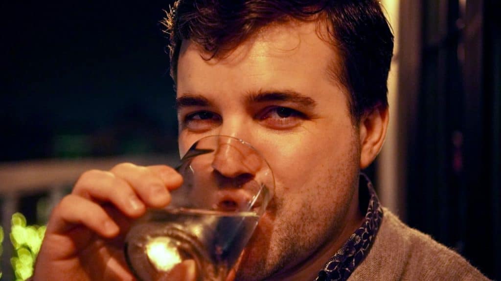 A man with dark hair looks at the camera while drinking from a glass.