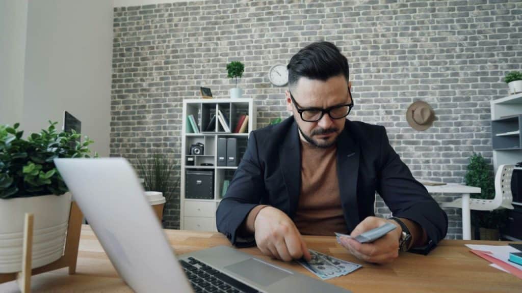 A man with a beard and glasses sits at a desk, counting money with a laptop in front of him.