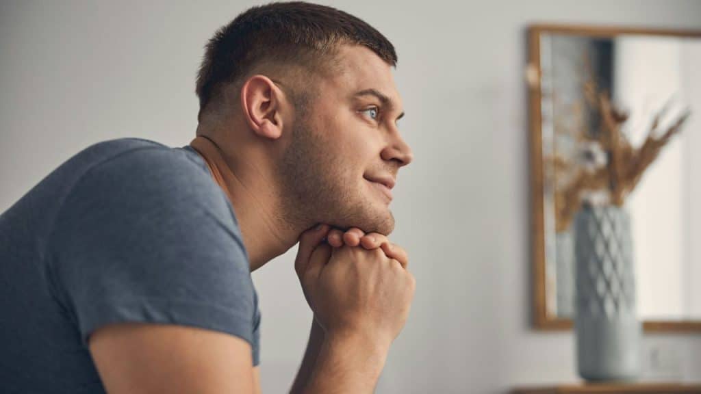 A man in a blue shirt with his hands clasped under his chin looks to the side.