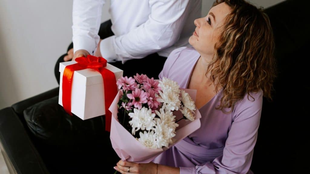 A woman holding a bouquet of flowers looks at a person who is giving her a gift box.