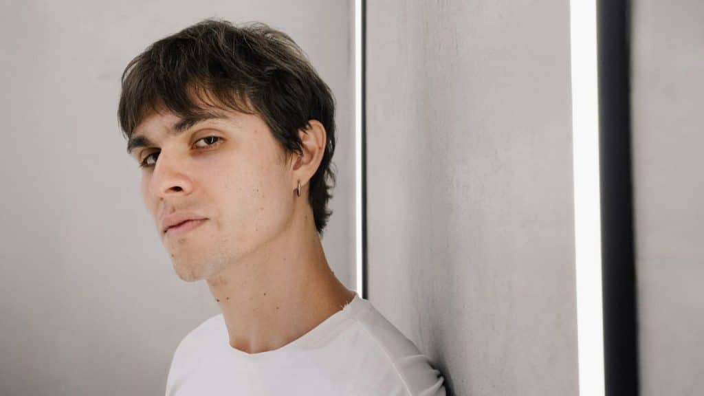 A man in a white shirt with a straight face leans against a wall.