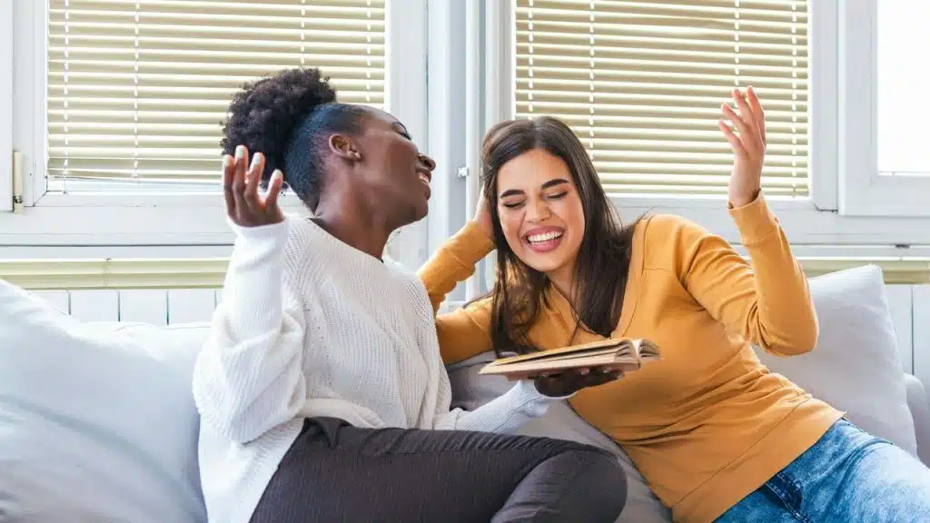 A pair of women laughing together on a couch with a book.