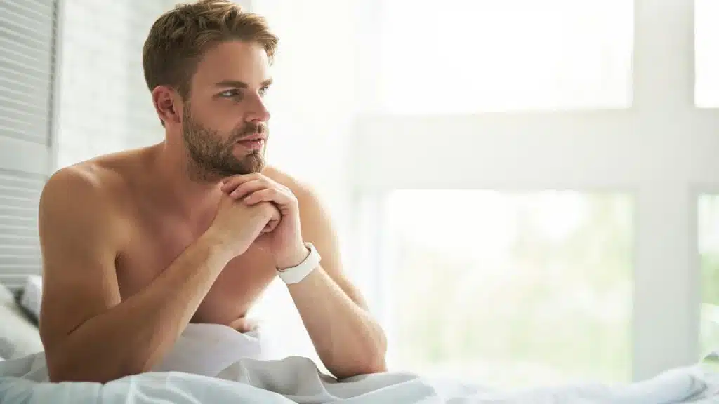 A shirtless man sitting in bed looking thoughtful.