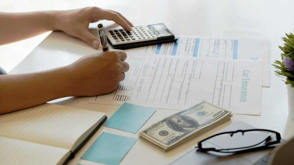 A person filling out car insurance forms with cash, a calculator, and a notebook on the desk.
