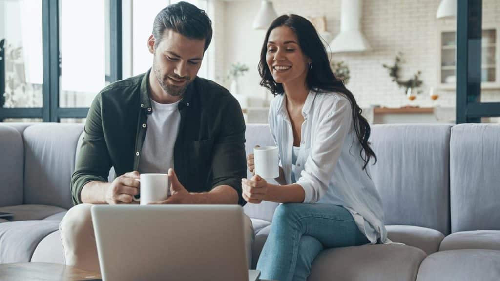 A couple sitting on a couch with coffee mugs, smiling while looking at a laptop.