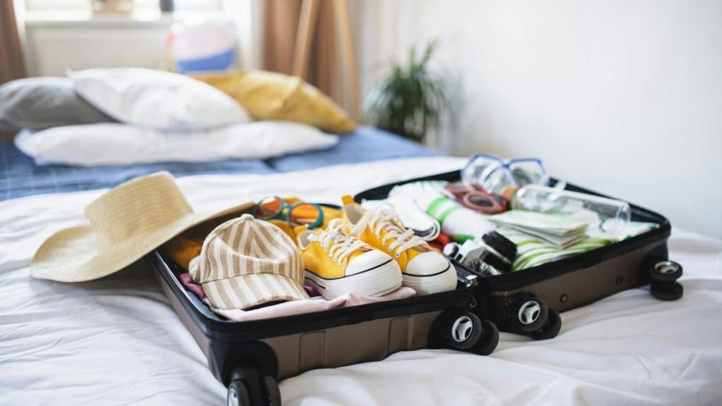 An open suitcase on a bed packed with clothes, shoes, hats, and travel essentials.