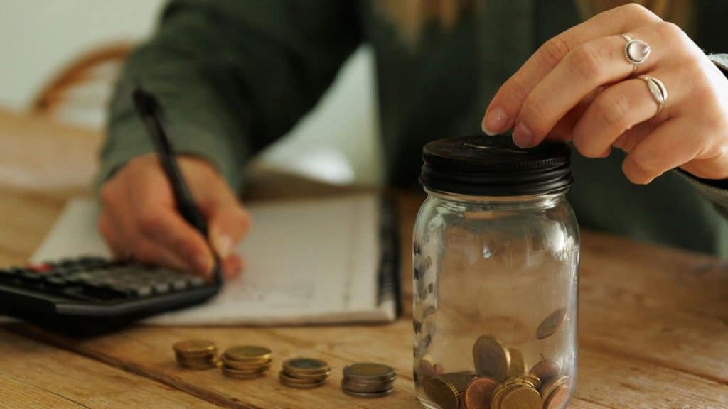 A person calculating finances while saving coins in a jar.