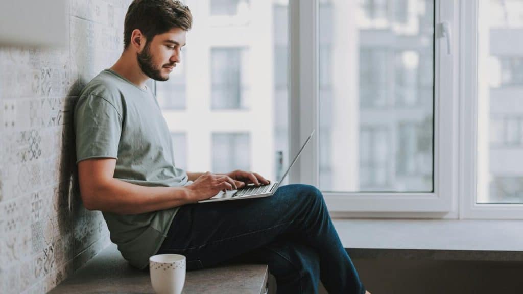A man sitting by a window working on a laptop with a coffee mug nearby.
