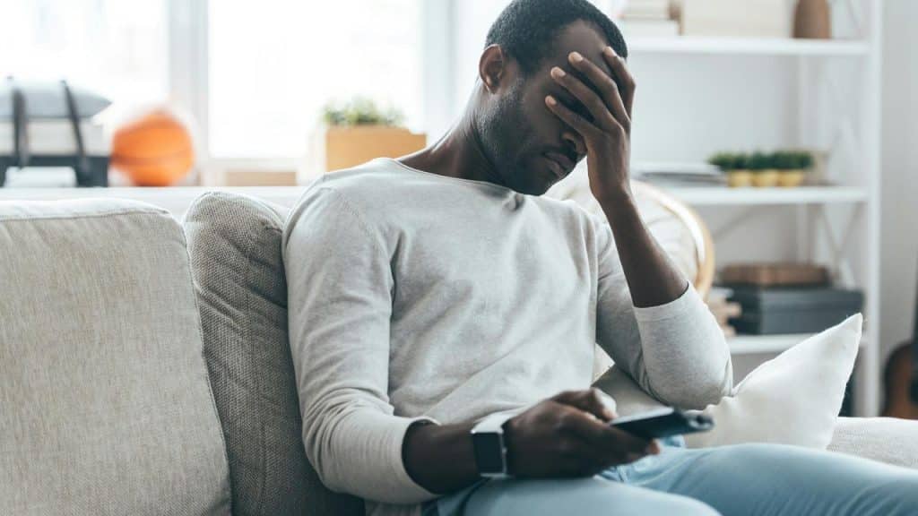 A man sitting on a couch holding a phone with his hand covering his face.