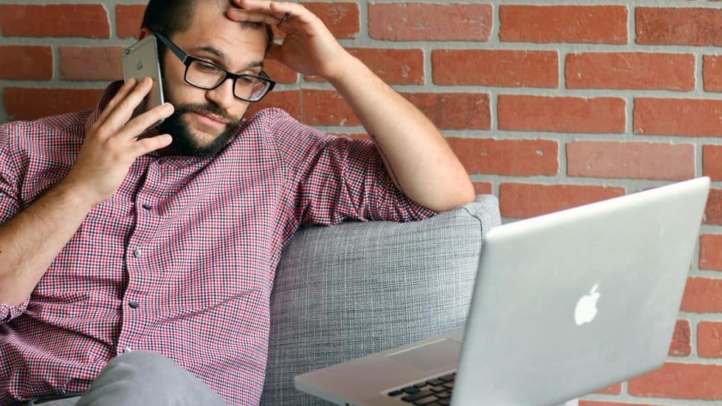A man talking on the phone while looking at a laptop with a concerned expression.
