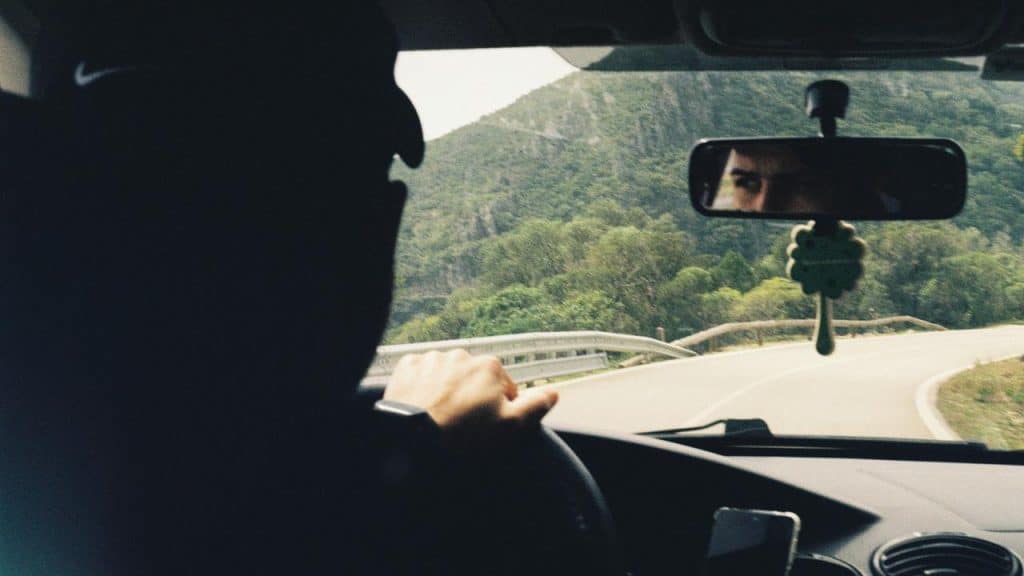 A person driving a car on a winding road with their eyes visible in the rearview mirror.