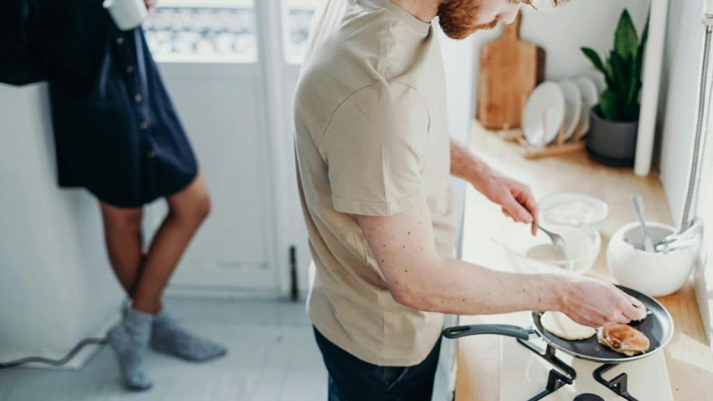A man cooking pancakes while a woman stands nearby with a mug.
