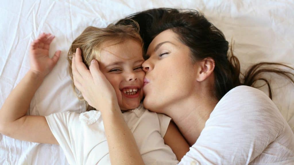 A mother kissing her laughing daughter while lying on a bed.