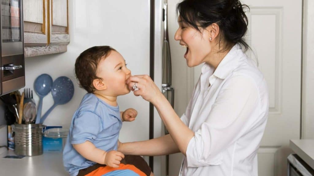A mother feeding her baby while smiling in the kitchen.