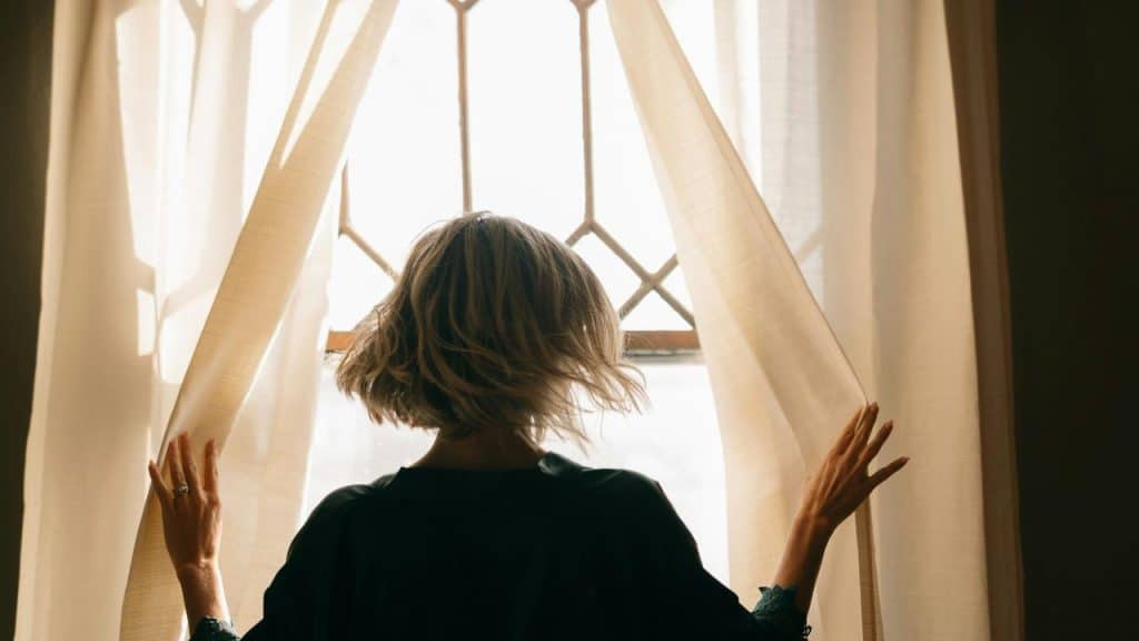 A woman opening curtains to let sunlight into the room.
