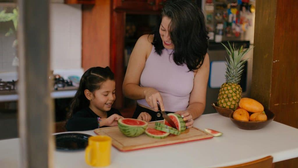 A mother cutting watermelon in the kitchen while her daughter smiles beside her.
