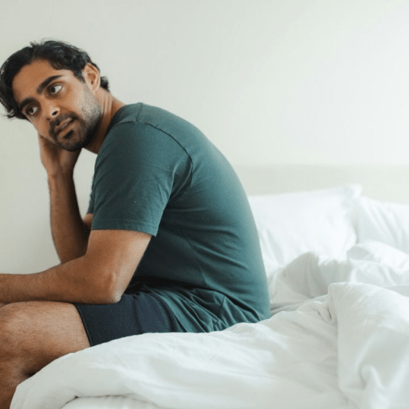 A pensive, bearded man in a dark green shirt sits on the edge of a bed.