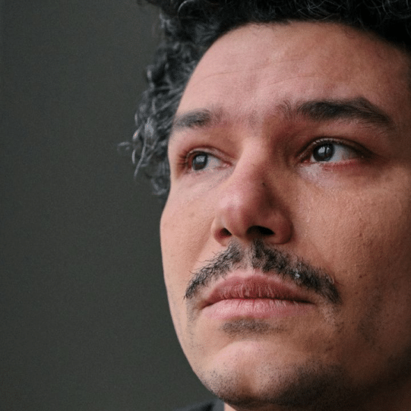 A sad man with a mustache and curly hair looks into the distance, with tears in his eyes.