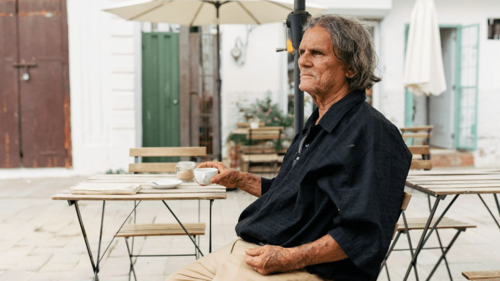 An elderly man sits at an outdoor cafe table, holding a small cup of coffee.