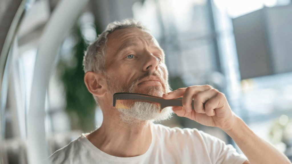A serious man with a gray beard and gray hair combs his beard in a mirror.