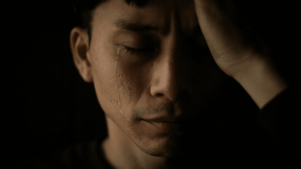 A crying man with his eyes closed, holding his forehead with his left hand.