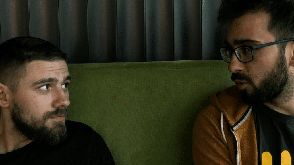 Two men with beards, one in glasses, look at each other seriously.