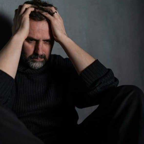 A sad, middle-aged man with a beard sits on the floor, holding his head in his hands..