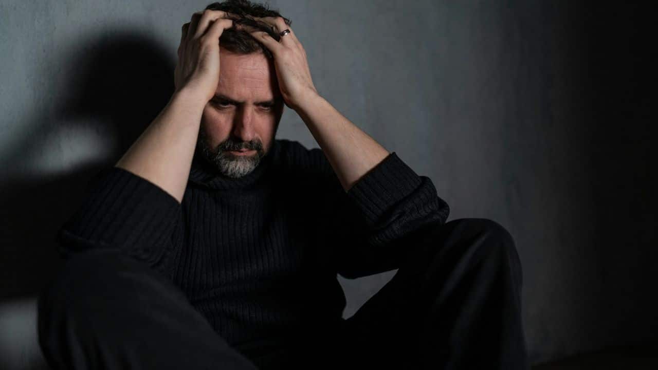 A sad, middle-aged man with a beard sits on the floor, holding his head in his hands..