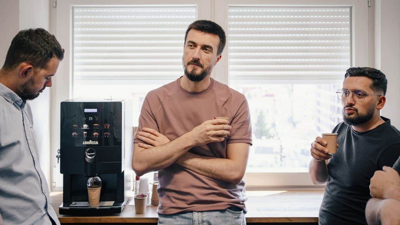 A handsome man with a beard and a pink shirt talks with his coworkers at a coffee machine.