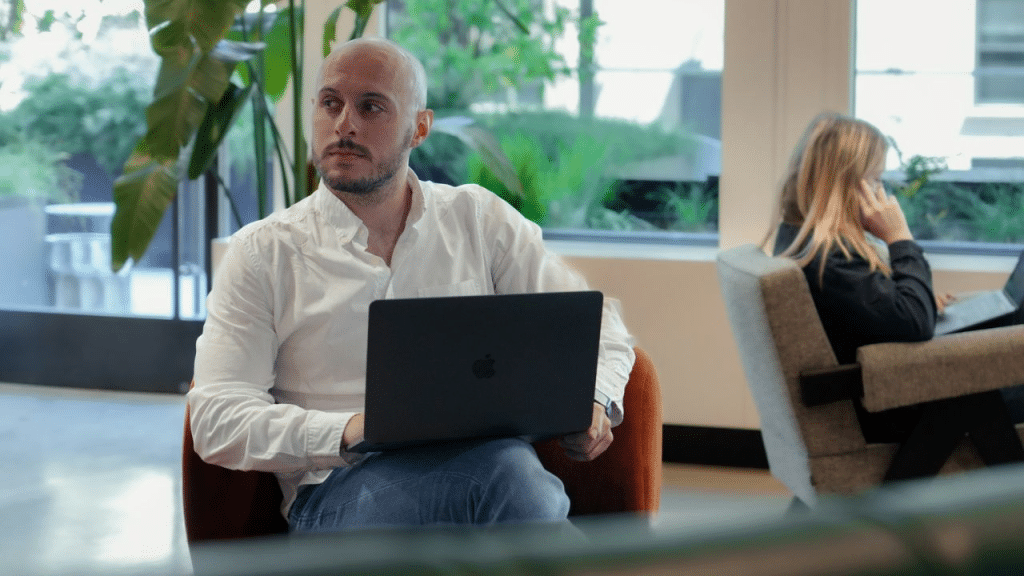 A thoughtful, balding man in a white shirt sits in a chair, holding a laptop. A woman sits in a chair behind him, talking on the phone.
