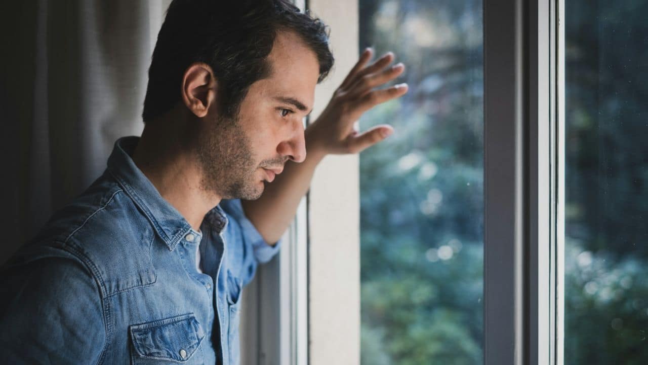 A worried man in a jean shirt stands alone and looks out a window.