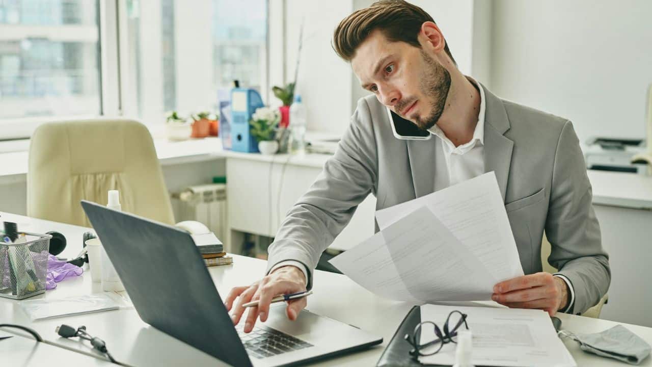 A busy businessman on the phone works on his laptop and holds some papers.