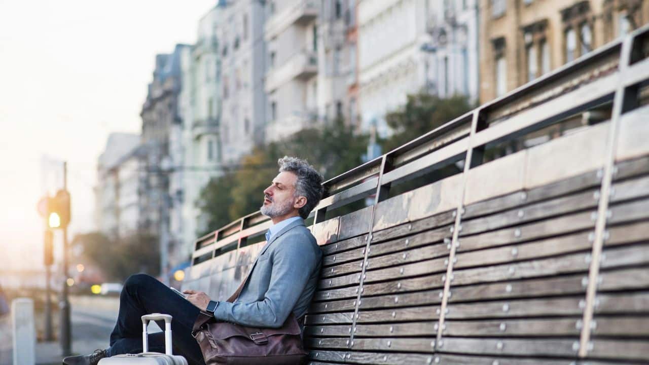 A middle-aged businessman with a beard sits on a bench, looking exhausted and sad.