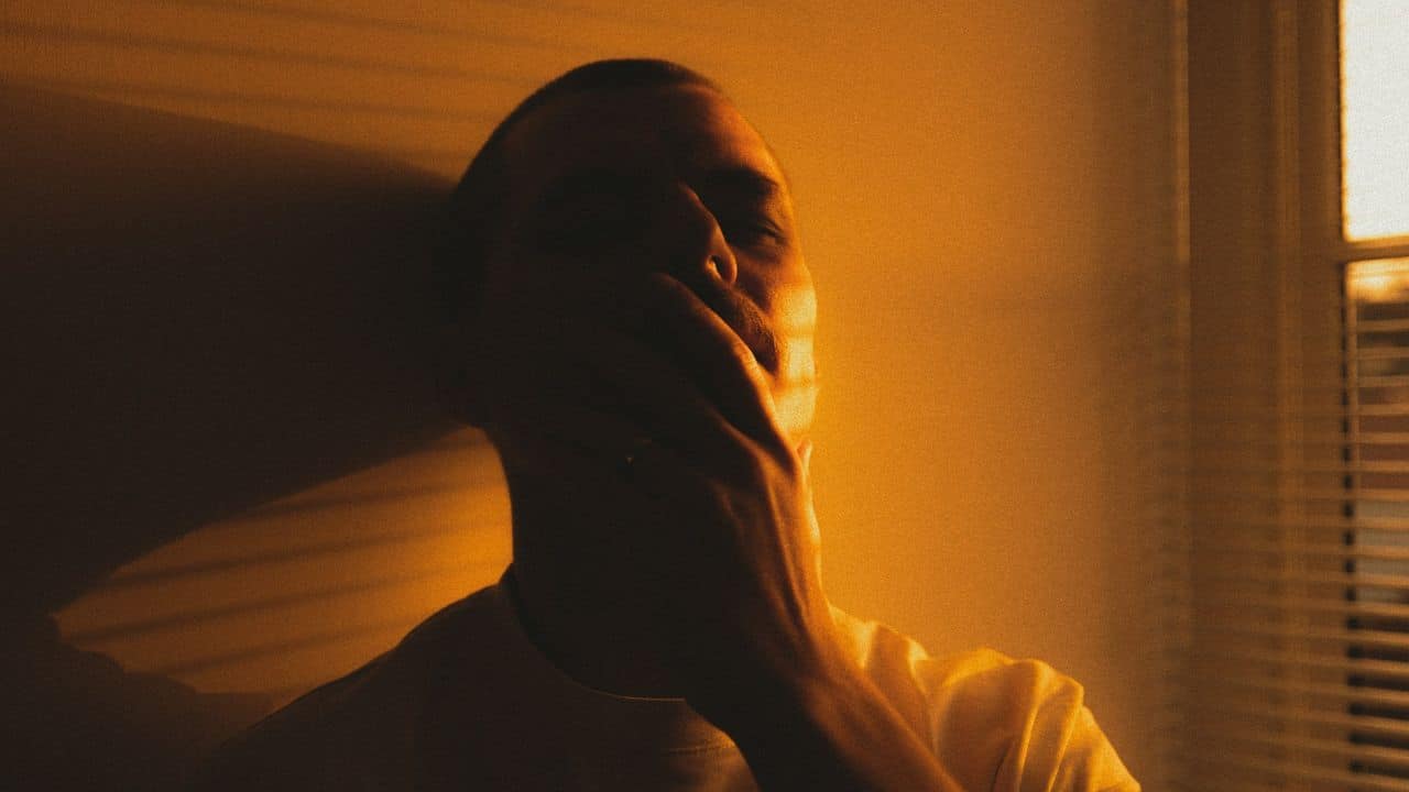 A man sits in the shadows, holding his hand over his mouth in sadness.