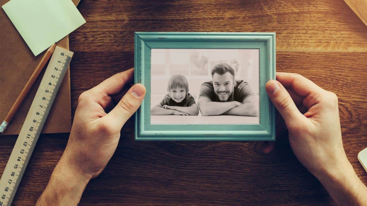 A pair of hands holds a framed black-and-white photo of a happy father and son.