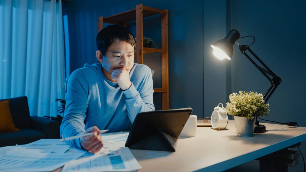 A frustrated man sits at a desk late at night, holding a pencil and looking at a tablet.