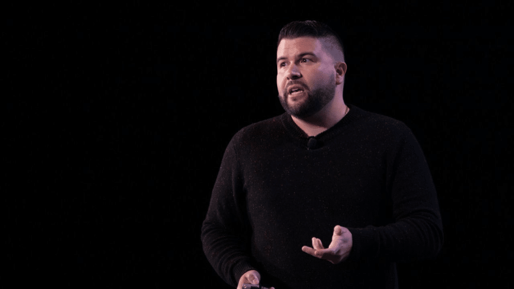 A man with a beard and dark hair stands against a black background, gesturing with his left hand as he talks.