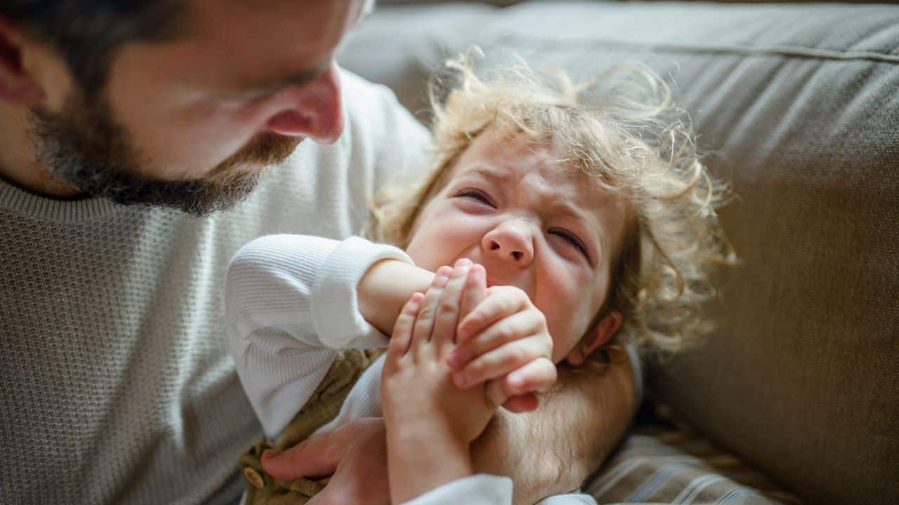 A distressed toddler with curly blonde hair cries and bites their hand while being held by a parent.
