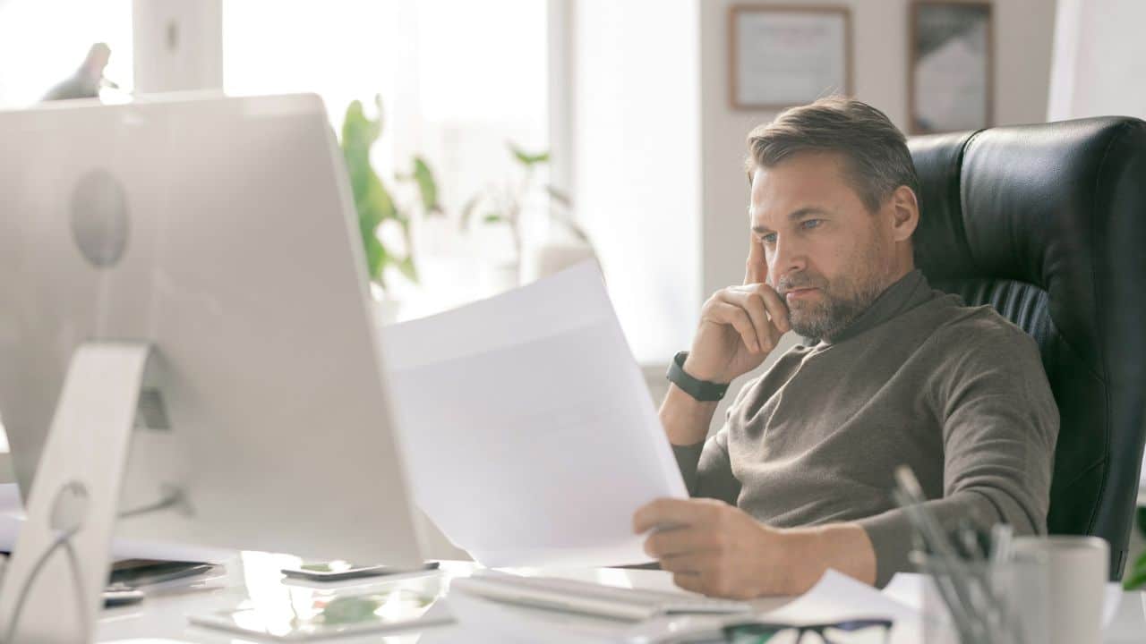 A handsome, middle-aged man with a beard looks at documents while sitting at his office desk.