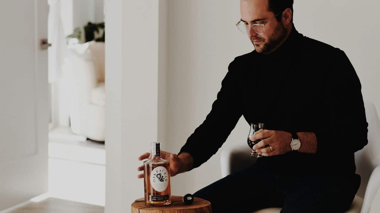 A man in a black shirt and glasses holds a bottle of whiskey and a glass.
