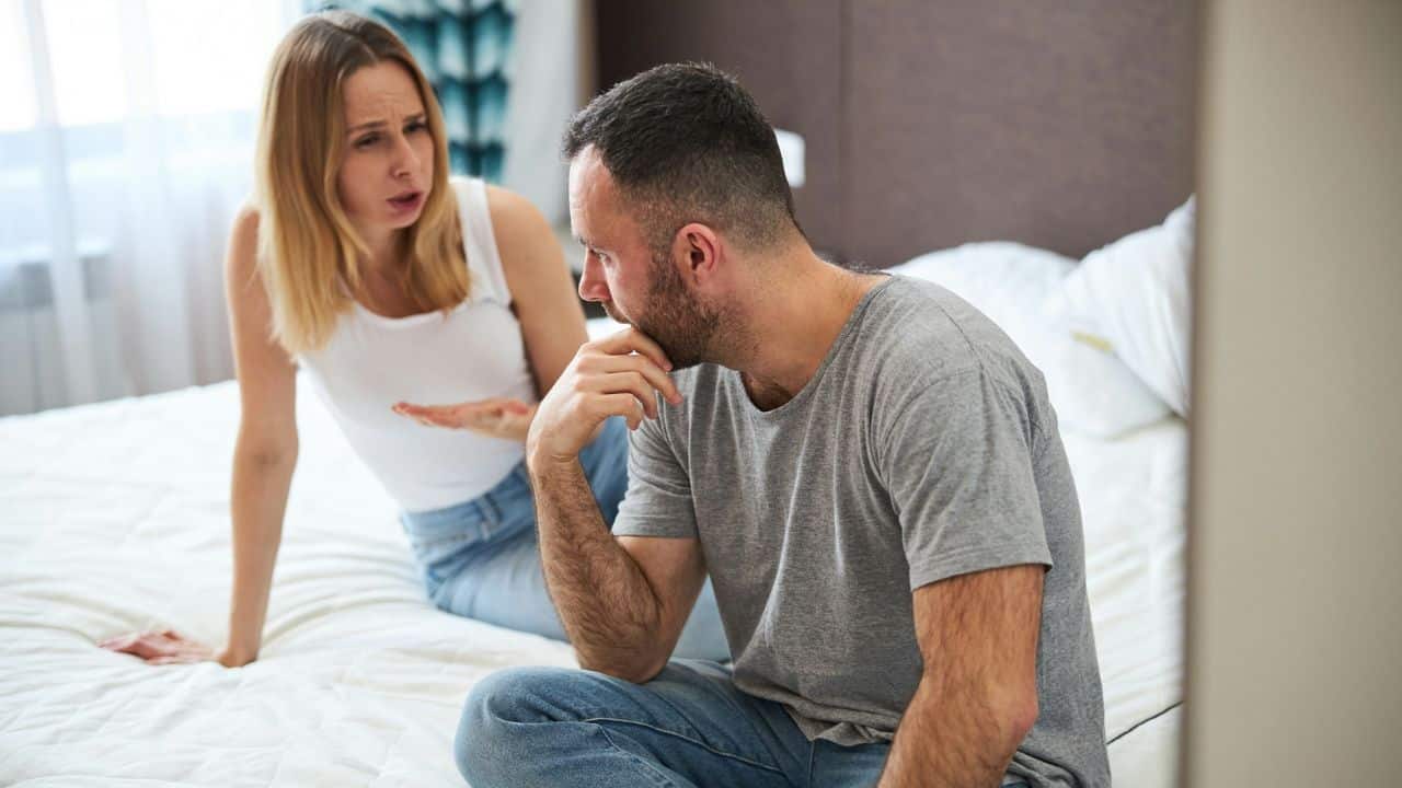 A worried man sits on a bed while a frustrated woman talks and gestures at him.