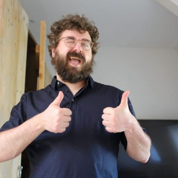 A happy, bearded man wearing glasses and a dark polo shirt gives two thumbs up.