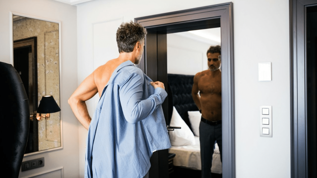 A man is putting on a blue shirt while looking at his reflection in a mirror.
